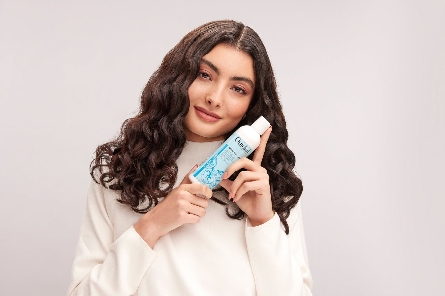 Model with loose brown curls holding a bottle of Moisture Lock® Leave-In Conditioner near their face
